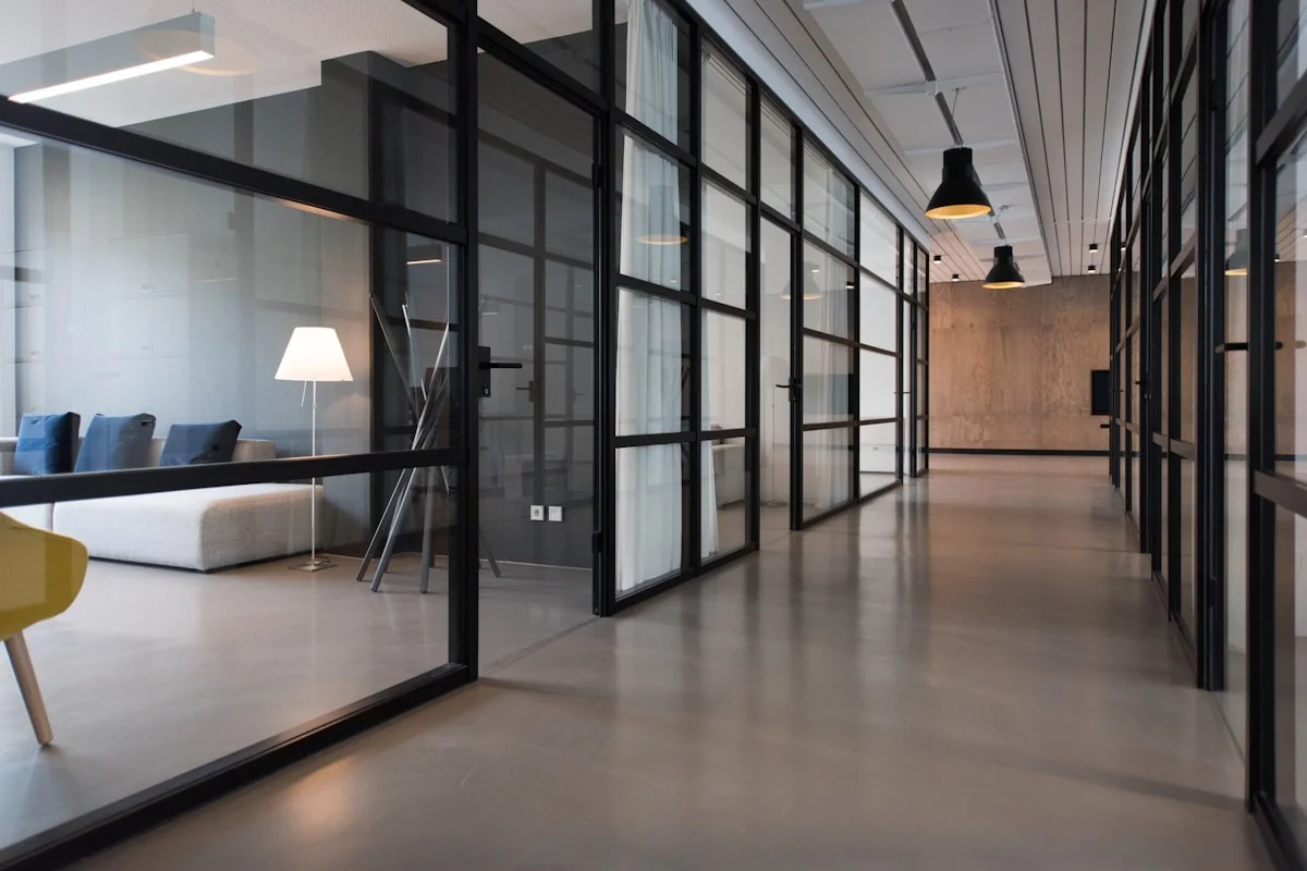 Modern glass-walled office corridor with lounge seating and pendant lights — professional workplace mood