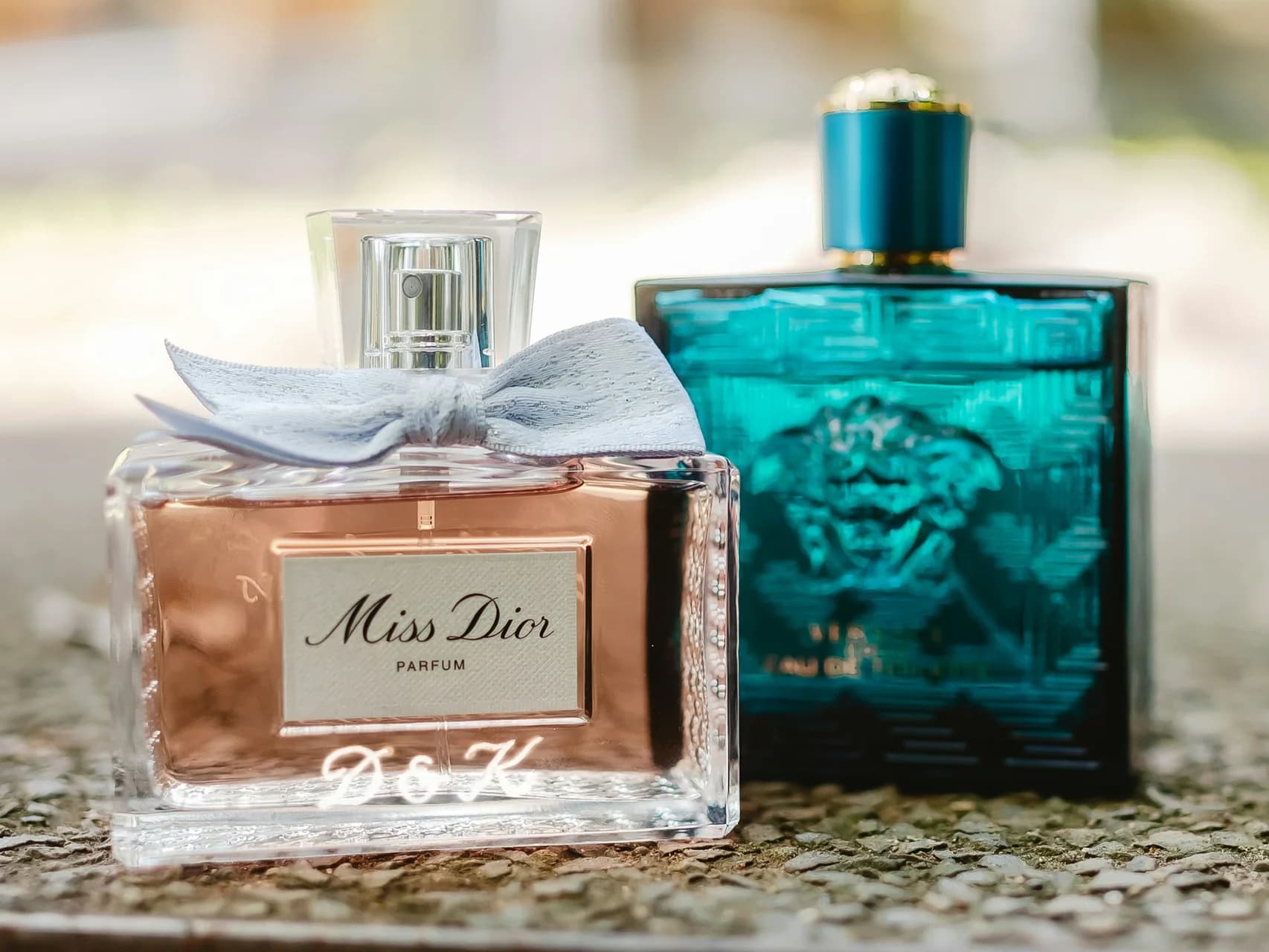Miss Dior bow-capped bottle beside teal Versace Eros bottle on stone surface — side-by-side longevity comparison still life